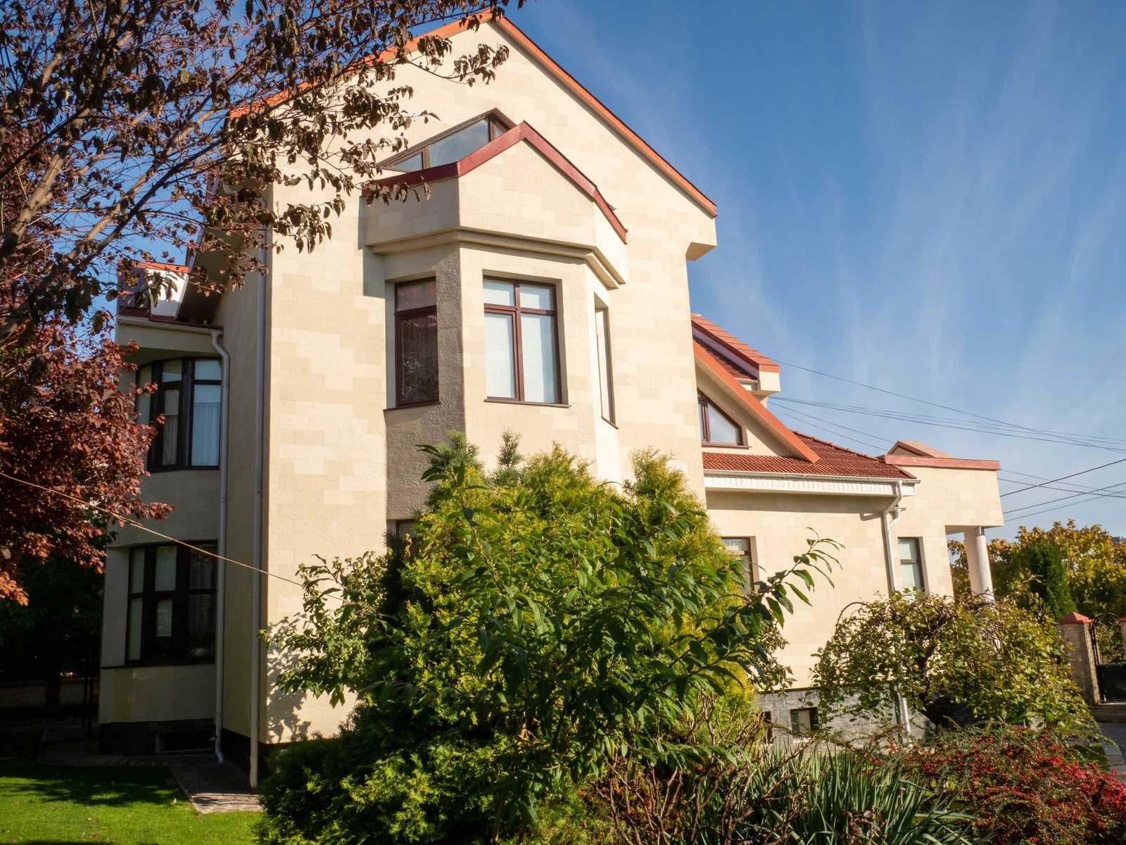 Casa văzută din grădină, cu bovindou, fronton înalt și fațadă din piatră flexibilă bej, printre arbuști