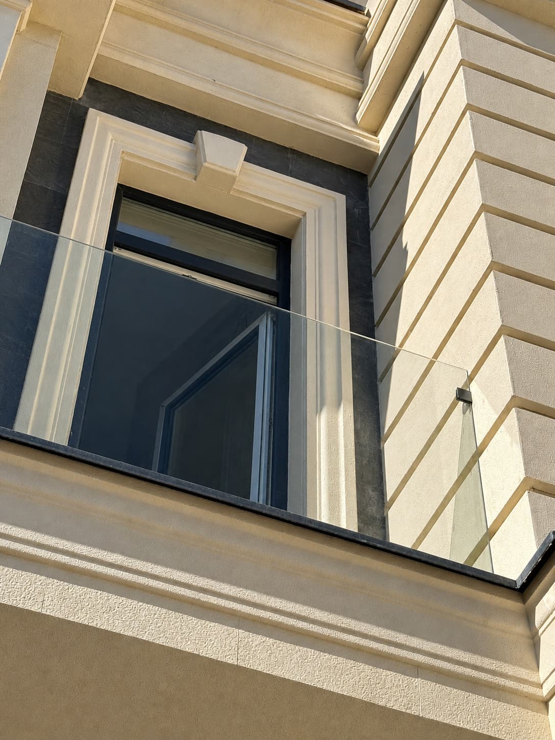 Fereastră cu balustradă de sticlă încadrată în piatră flexibilă bej, deasupra unei cornișe pe console