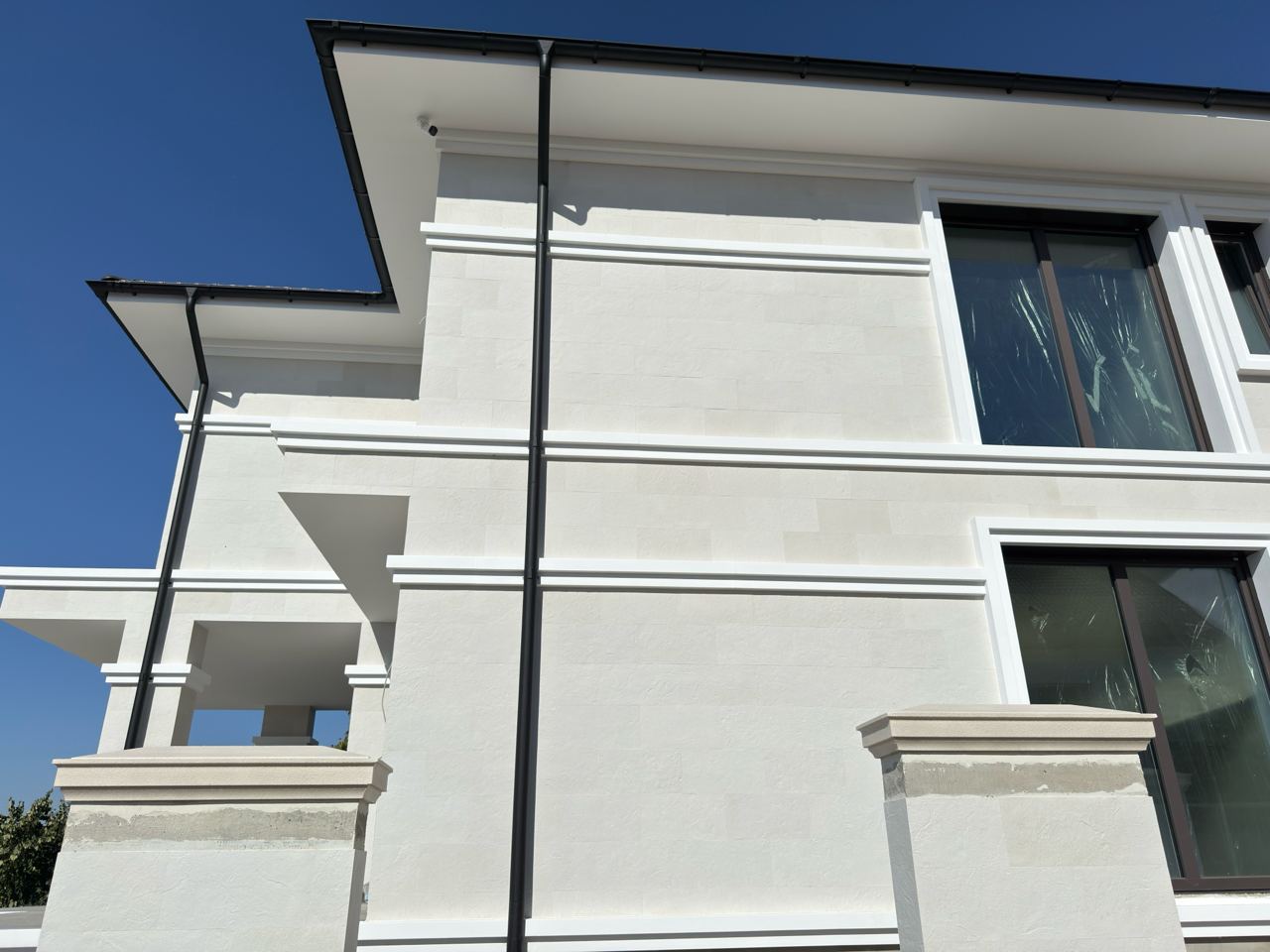 Colțul casei dinspre grădină, cu balcon, cornișe albe și burlan negru pe piatra flexibilă